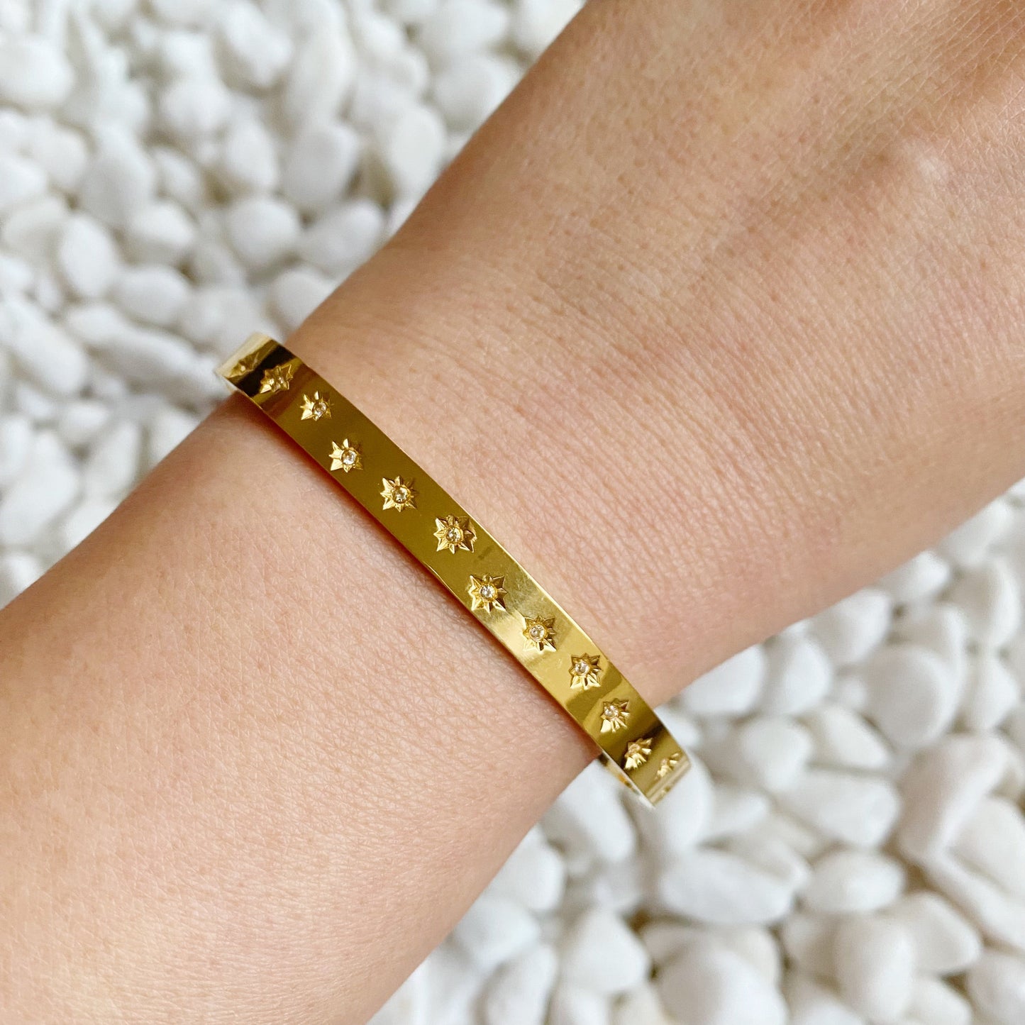 A person's wrist wearing a golden bracelet with shiny stars around, placed on a surface with white decorative rocks.