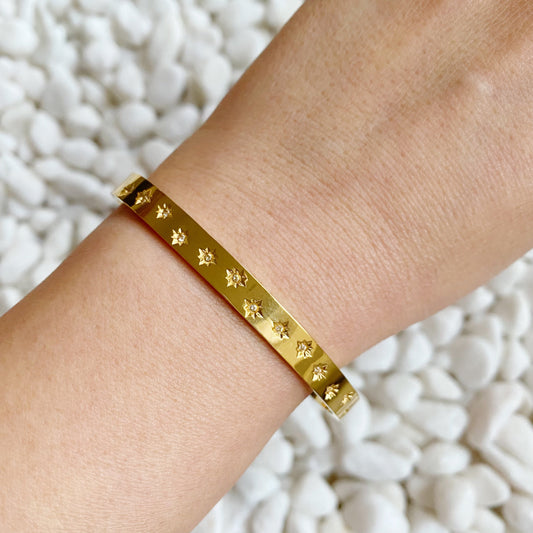 A person's wrist wearing a golden bracelet with shiny stars around, placed on a surface with white decorative rocks.