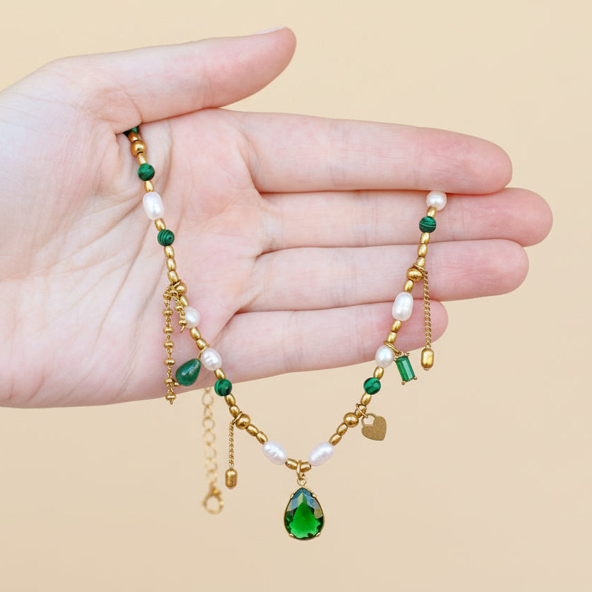 Hand holding a delicate necklace with green and gold elements on a beige background